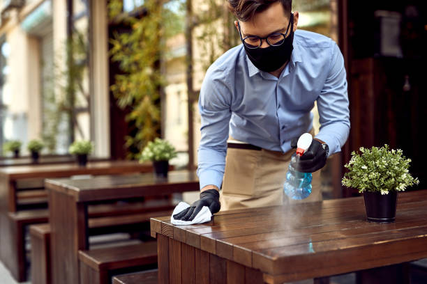 Waiter wearing protective face mask while disinfecting tables at outdoor cafe.