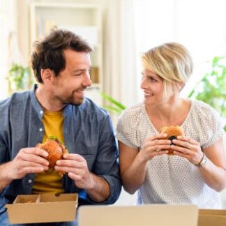 Front view of happy couple sitting on sofa indoors at home, eating hamburgers.