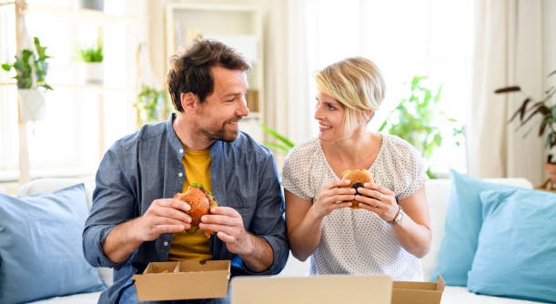 Front view of happy couple sitting on sofa indoors at home, eating hamburgers.